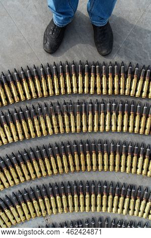 Lots Of Bullets For A Heavy Machine Gun Lie On The Asphalt Near The Feet Of A Man In Civilian Clothe