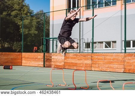 Blond Boy In Sportswear Jumps Over Red Obstacles To Improve Lower Body Dynamics. Plyometric Training