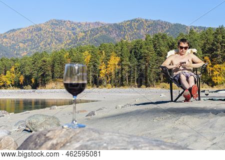 Fashionable Attractive Man, With Naked Torso Sits In Chair On Beach In Sunglasses, Looks Smartphone,