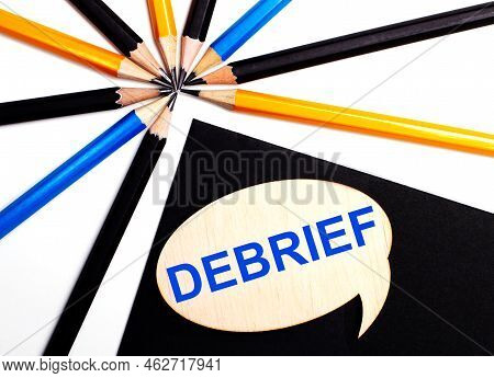Wooden Card With The Text Debrief On A Black Background Near Multicolored Pencils.