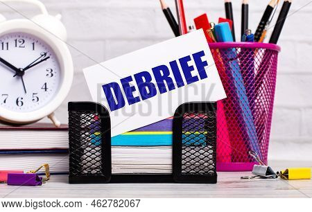 The Office Desk Has Diaries, An Alarm Clock, Stationery, And A White Card With The Text Debrief. Bus
