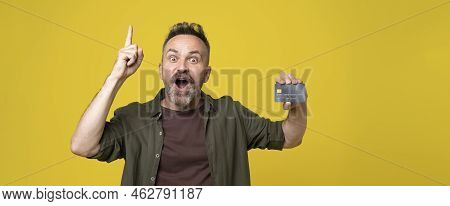 Astonished Mature Man Notes With Pointing Up Hand Hoding Bank Card Isolated On Yellow Background, St