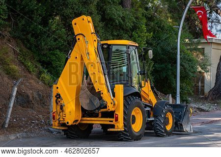 Yellow Wheeled Excavator Standing On The Side Of The Road. The Turkish Flag Flies On A Pole.