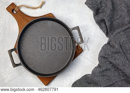 Black Cast Iron Empty Frying Pan On A Wooden Cutting Board And A Gray Kitchen Towel On A Gray Concre