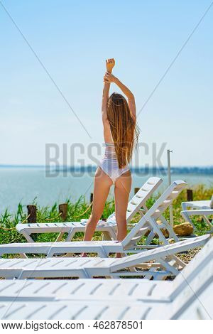 A Long-haired, Slender, Beautiful Woman In A Swimsuit By A Chaise Longue By Sea.