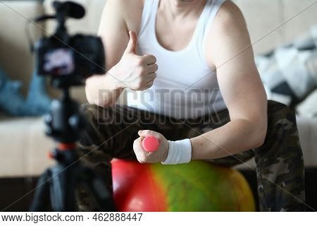 Athlete Man Holds Thumbs Up And Raises Dumbbells Sitting On Ball At Home In Front Of Camera. Recomme
