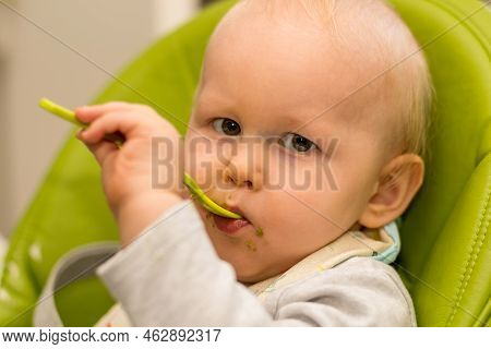 Little Baby Playing And Feeding Himself With Spoon Sitting In Highchair. Baby First Solids. Weaning.