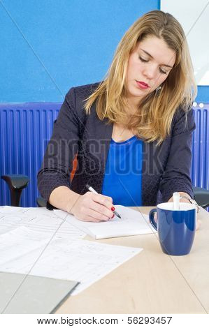 Young attractive woman concetrated at work, working on a technical design of a product whilst taking notes behind her desk
