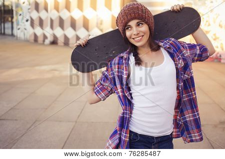 Brunette teenage girl in hipster outfit (jeans shorts keds plaid shirt hat) with a skateboard at the