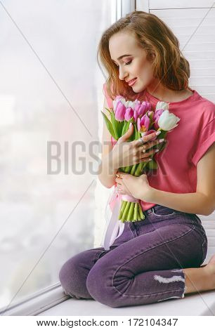Pretty young woman in pink T-shirt with bouquet of tulips on window-sill