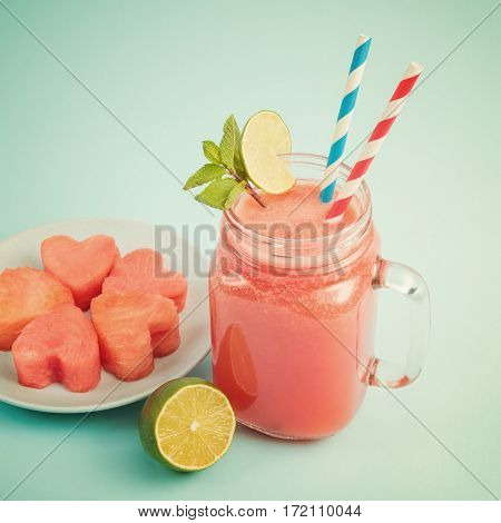 Close-up of watermelon smoothie in Mason jar decorated with lime, mint, straws. Watermelon slices curved like hearts on plate