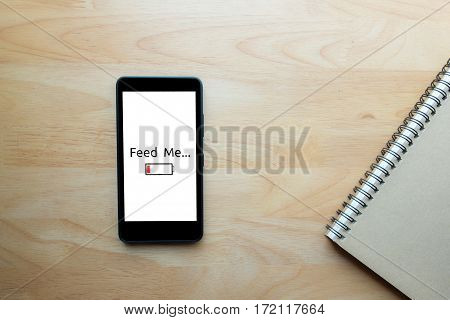 Smartphone show text on screen "feed me" status show low battery with notebook on wooden table background.