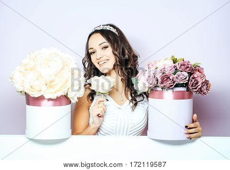 Sexy young pretty woman or cute smiling girl with crown on head has long hair in white dress with lilac rose and peony flowers bouquet boxes on violet background