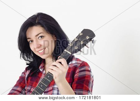 Young cute girl in a checkered shirt holding an acoustic guitar and smiling.