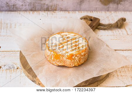 Whole wheel of soft French,German cheese with orange rind with mold on parchment paper, wood cutting board, concrete wall rustic kitchen interior