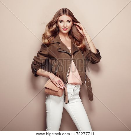 Young blond woman in brown jacket and white jeans. Girl posing on a beige background. Jewelry and hairstyle. Girl with brown handbag. Fashion photo