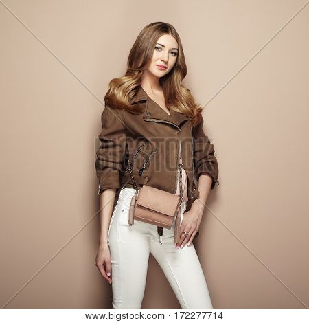 Young blond woman in brown jacket and white jeans. Girl posing on a beige background. Jewelry and hairstyle. Girl with brown handbag. Fashion photo