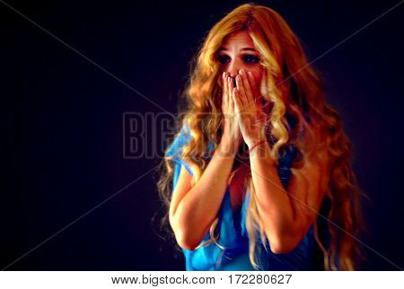 Frightened woman screaming with fear indoor at halloween night. Horror. Worried girl with tousled hair worried bawl and l covers her mouth with her hands black background.