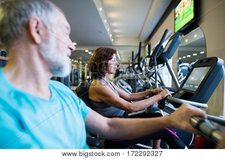 Beautiful fit senior couple in sports clothing in gym doing cardio workout, exercising on recumbent bicycle. Sport fitness and healthy lifestyle concept.