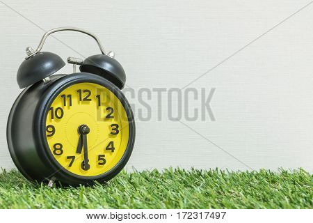 Closeup black and yellow alarm clock for decorate show half past six or 6:30 a.m. on green artificial grass floor and cream wallpaper textured background with copy space