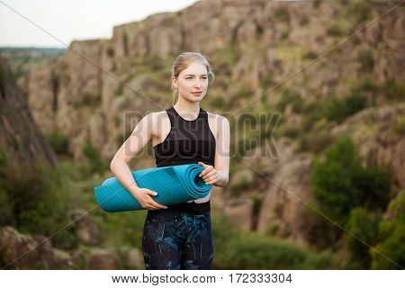 Young sportive girl smiling, holding yogamat, standing on rock in canyon. Copy space.