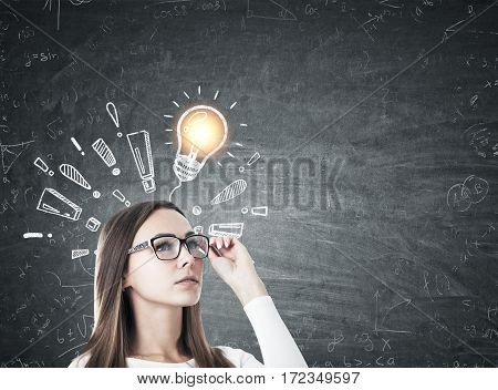 Portrait of a serious businesswoman in glasses standing near a blackboard with a light bulb sketch surrounded by exclamation marks. Mock up.