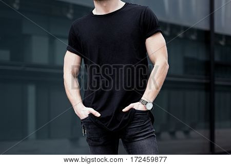 Young caucasian muscular man wearing black tshirt and jeans posing in center of modern city. Blurred background. Hotizontal mockup