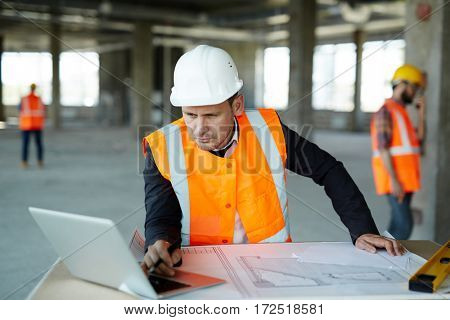 Portrait of development supervisor wearing protective vest and helmet over formal suit  proofing blueprints using laptop computer inside unfinished building