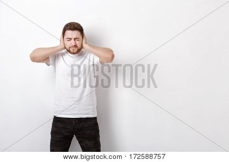 portrait of a young Brunet man in white shirt covers his ears with his hands against a grey background. I don't want to hear anything