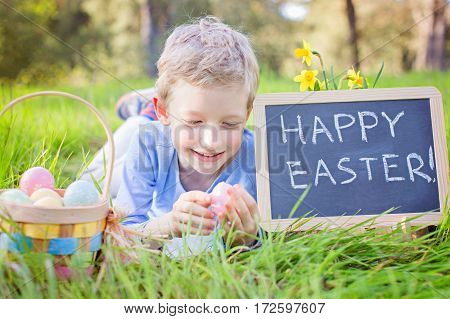 smiling positive kid lying with basket full of easter eggs after egg hunt and blackboard sign saying "happy easter!" enjoying blooming time at spring in the park