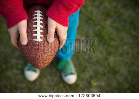 Top view on hands of little kid boy holding football on the stadium on a sunny day. Child ready to throw a football. Sport concept. Sport activities for children outdoors.