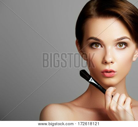 Beautiful Young Woman Applying Foundation On Her Face With A Make Up Brush Isolated On Gray Backgrou