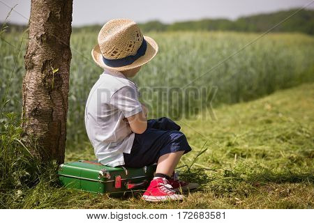 Cute little kid boy in shorts white polo and straw hat sits on his green suitcase in a field near a tree waiting for a bus. summer portrait of a small child in profile retro style. Ready to travel.