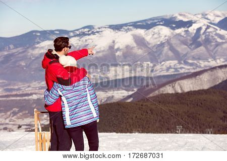 Rear view of a loving couple in fur hood jackets looking at snowed mountain range.