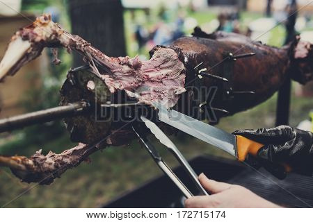 Barbecue picnic at country fair. Lamb roasted at spit. Grill, big piece of meat at rolling skewer. Street vendor, professional chef hands cooking bbq food, unrecognizable. Catering concept