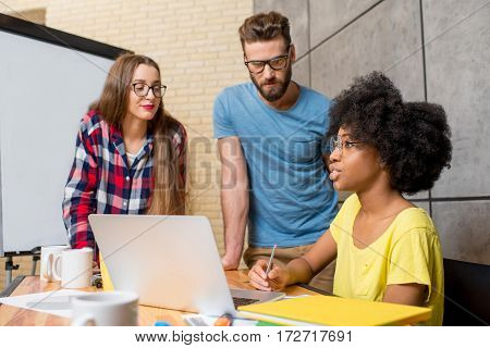 Multi ethnic coworkers dressed casually in colorful clothes talking together sitting with laptop indoors