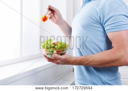 Athletic man eat healthy lunch in modern interior. Unrecognizable profile male torso in blue t-shirt, hand with fork, near window with vegetable salad in bowl, diet food concept. High key image