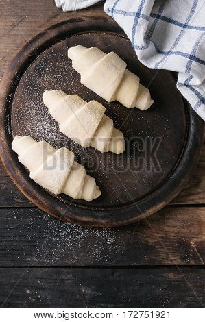 Raw unbaked croissant on wooden chopping board with kitchen towel over old dark wood background. Top view