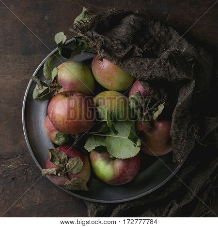 Vintage metal plate with harvest of wild apples with leaves on sackcloth rag over old wooden background. Top view, dark rustic style