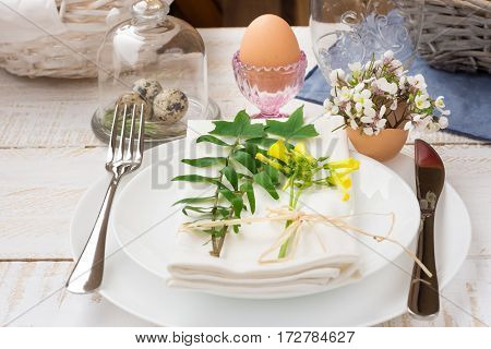Easter table setting white plates cutlery napkin white flowers in eggshell green twigs crystal cup basket outdoors kinfolk wood