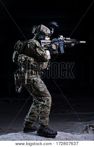 Soldier with rifle in helmet with night vision device aiming to target on dark background