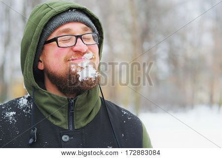 Portrait of young smiling man in glasses with a snowy beard during snowfall in winter Park closeup