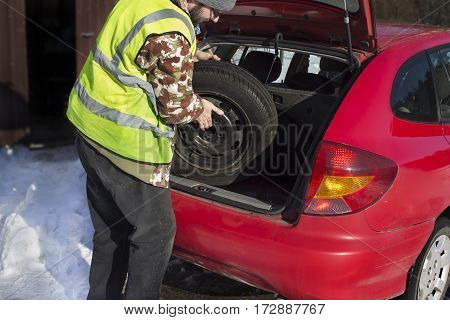 The man pulls out a spare wheel from the trunk of a car wearing a reflective vest. Replacing the spare wheel in winter conditions.