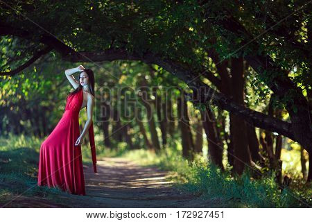 Wonderful sexy fashion model walking in a fantastical forest by footpath. Photo of sensual seductive woman in full length in luxury long red dress posing against bokeh trees background. Multi-racial Asian Caucasian girl. Fashionable toning. Creative compu