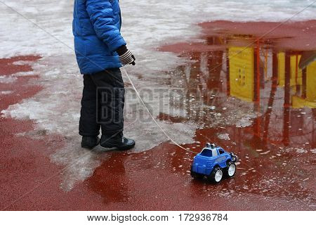 Spring. The snow is melting. The image of a child dressed in a jacket boots gloves. A child plays on the Playground with toy car: the car driven through a puddle on the rope.