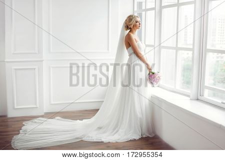 Classical young gourgeous bride. Studio interior fashion shot of fashion model in wedding dress with bouquet of flowers in white room with big window. Blonde woman portait with profeshional make-up and hairstyle.