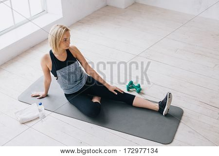 Attractive slim female doing stretching exercises on black mat in modern bright fitness center. She has some rest after hard fitness exercise with dumbbells