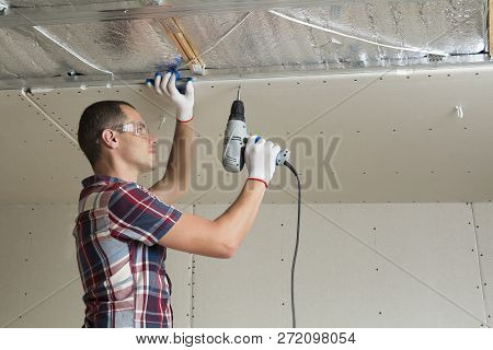 Young Man In Goggles Fixing Drywall Suspended Ceiling To Metal Frame Using Electrical Screwdriver On
