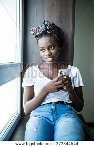 Young African Woman Typing On Mobile Phone On Windowsill At Home