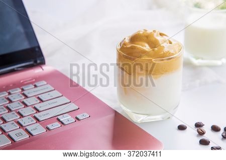 Close Up A Cup Of Dalgona Coffee With Coffee Beans Next To A Pink Laptop On A White Table With A Cup
